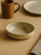 Płytka, nakrapiana beżowa miska ceramiczna stoi na jasnym drewnianym stole. Minimalistyczna aranżacja naczyń obejmuje brązowy kubek, pasujący talerz oraz lnianą serwetkę w tle.