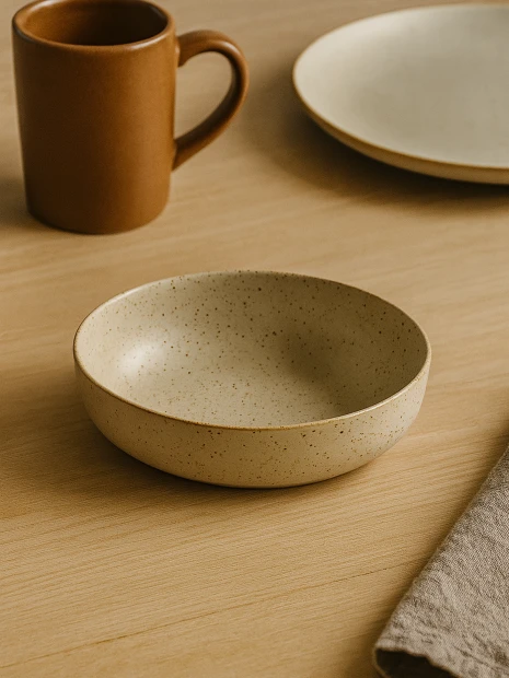 A shallow, speckled beige ceramic bowl is placed on a light wooden table. The minimalist tableware arrangement includes a brown mug, a matching plate, and a linen napkin in the background.